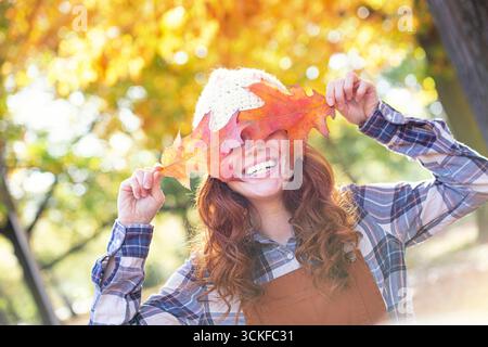 donna dai capelli rossi che indossa un berretto che tiene le foglie autunnali davanti agli occhi e sorride alla fotocamera Foto Stock