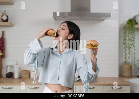 Sorridente giovane donna in piedi in cucina che mangia deliziosi hamburger tenendo in mano un altro panino, simboleggiando fast food, stile di vita, appetito, sn Foto Stock