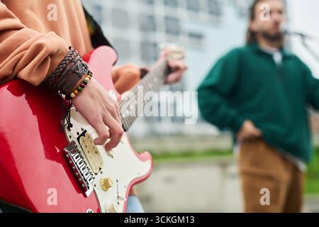 Giovane donna caucasica adulta che suona la chitarra elettrica all'aperto con un giovane uomo caucasico adulto che canta in sottofondo, concentrandosi sulla mano che canta le corde durante le esibizioni dal vivo Foto Stock