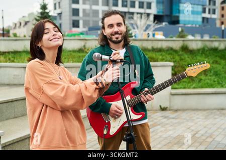 Giovane donna caucasica sorridente e tenendo il microfono mentre si trova accanto al giovane caucasico che suona la chitarra elettrica all'aperto, entrambi guardando verso la telecamera Foto Stock