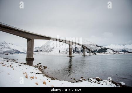 Un ponte curvo si estende su un fiume innevato con montagne sullo sfondo. Foto Stock