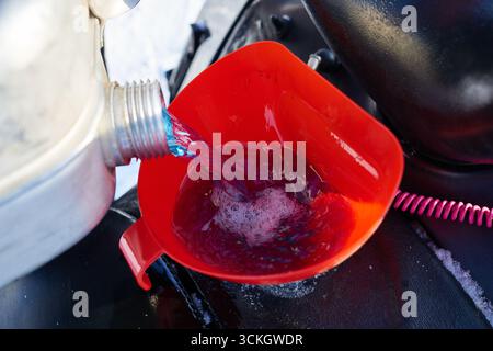 Primo piano che mostra un uomo sconosciuto che versa liquido di raffreddamento liquido blu dal contenitore di plastica nell'imbuto rosso, riempiendo il vano motore del veicolo durante la manutenzione invernale all'aperto Foto Stock