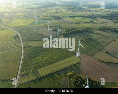 Vista aerea delle turbine eoliche alte contro il mosaico di campi verdeggianti e la luce del sole dorata, Fayl-Billot, Grand Est, Francia. Foto Stock