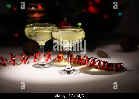 Due eleganti bicchieri pieni di bevande frizzanti siedono su un tavolo, decorati con nastri luminosi e ornamenti natalizi sullo sfondo, che evocano un'atmosfera celebrativa durante un incontro festoso. Foto Stock