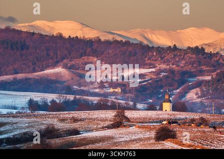 Vista aerea di un paesaggio sereno con una chiesa che cammina all'orizzonte, annidata tra colline innevate e montagne lontane baciate dal sole, Oravce, regione di Banská Bystrica, Slovacchia. Foto Stock