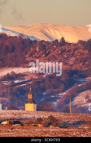 Vista aerea della pittoresca guglia della chiesa che attraversa il paesaggio invernale, con campi polverati di neve che conducono a montagne lontane baciate dal sole, Oravce, Banská Foto Stock