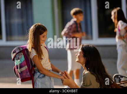 La gioiosa riunione si svolge quando la madre abbraccia sua figlia dopo la scuola. Altri bambini giocano e socializzano in sottofondo nelle giornate di sole Foto Stock