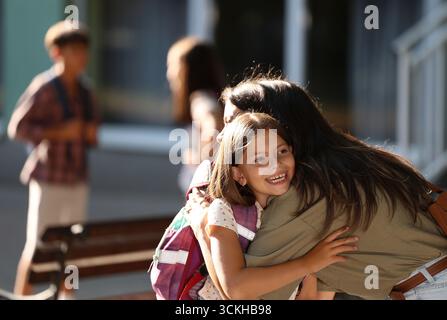 La gioiosa riunione ha luogo quando la madre abbraccia sua figlia dopo la scuola. Altri bambini giocano e socializzano in sottofondo nelle giornate di sole Foto Stock