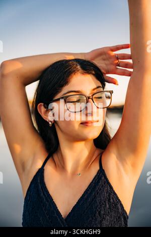 Giovane donna che indossa gli occhiali che si rilassa con le braccia al tramonto Foto Stock