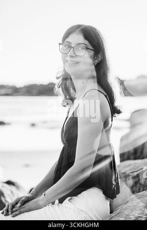 Giovane donna che indossa occhiali sorridenti in spiaggia durante l'ora d'oro Foto Stock