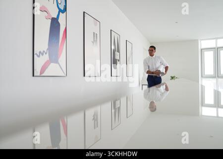 Giovane adulto in piedi in un moderno corridoio d'ufficio, guardando opere d'arte astratte incorniciate sulle pareti, interni luminosi e minimalisti con grandi finestre Foto Stock