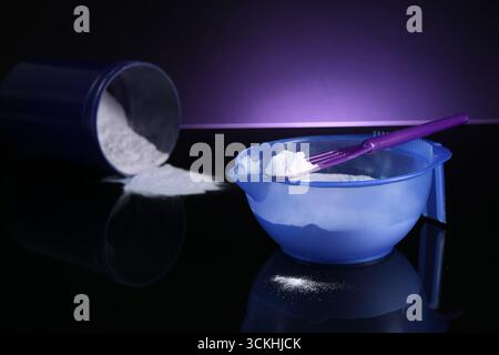 Prodotto cosmetico biondo per la cura dei capelli. Polvere nel recipiente, nel recipiente e nella spatola sulla superficie a specchio nera su sfondo viola scuro, primo piano Foto Stock