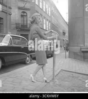 Nel 1956, quando i parchimetri richiedevano ancora monete, una giovane donna parcheggia la sua auto in una strada trafficata e inserisce le monete nel parcheggio accanto al suo posto auto. Dopo aver inserito le monete, gira il quadrante per impostare il tempo di parcheggio a pagamento e l'orologio del contatore inizia il conto alla rovescia. Questa scena cattura il fascino della vita urbana della metà del XX secolo, con auto d'epoca e contatori meccanici che riflettono un'era più semplice e basata sul denaro. Kristoffersson rif. BZ28-2. Foto Stock