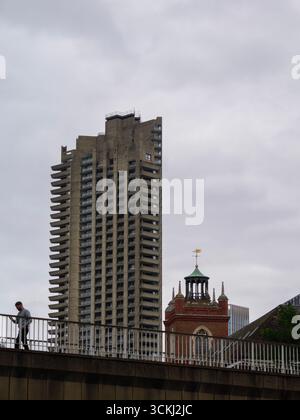 Barbican, City of London, Regno Unito. Pedoni che camminano lungo la passeggiata Barbican Highwalk con una torre residenziale nel centro e la chiesa di St Giles-without-Cripplegate, classificata di II grado, e la guglia visibile sulla destra Foto Stock