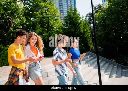 Gruppo di giovani amici che si divertono insieme all'aperto durante il giorno, utilizzando gli smartphone e sorridendo mentre camminano Foto Stock