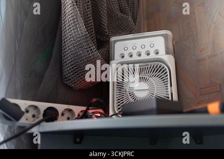 Ventola elettrica portatile su ripiano con striscia di alimentazione. Piccola ventola elettrica su un ripiano accanto a una presa multipla, simbolo del comfort domestico, del consumo energetico e della vita moderna Foto Stock