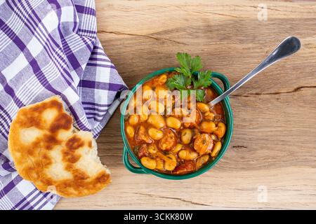 Stufato di fagioli di burro con salsa di pomodoro e salsicce di maiale a fette, pane pita e un asciugamano viola su un tavolo in legno di quercia Foto Stock