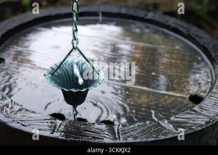 La coppa della catena antipioggia, in metallo, è appesa su un barile di legno. La tazza ha un bordo smerlato e una patina color ottanio, sospesa da maglie intrecciate. L'acqua scorre verso il basso Foto Stock