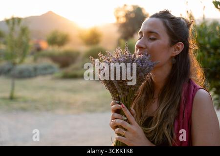 Giovane donna che profuma un bouquet di fiori di lavanda in un campo al tramonto, godendo la fragranza e la luce dorata dell'ora Foto Stock