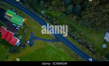 Immagine aerea di un'area rurale in Islanda caratterizzata da una strada pavimentata, edifici colorati con tetti rossi e verdi, alberi fitti e un piccolo ruscello. Foto Stock