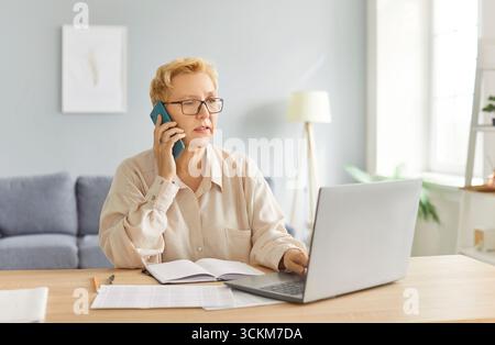 Donna anziana seduta alla scrivania con laptop e documenti finanziari e che parla tramite telefono cellulare Foto Stock