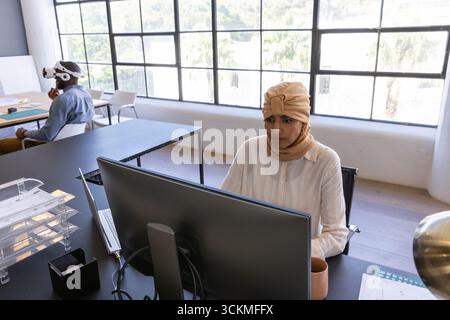 Diversi colleghi che indossano un velo abbronzato e un visore vr che collaborano al computer in ufficio Foto Stock