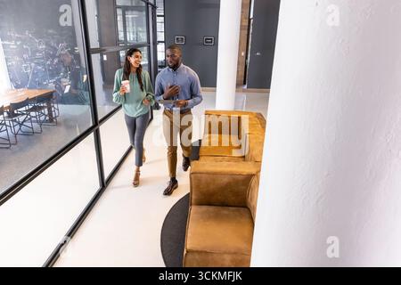 Diversi colleghi che camminano nella hall dell'ufficio oltre la parete in vetro, divano in pelle con tazza da caffè, smartphone Foto Stock