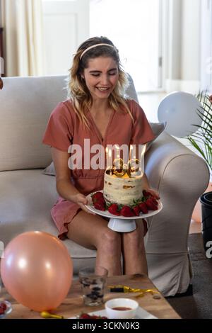 Donna seduta sul divano di casa che tiene una torta di compleanno con il numero 30 di candele accese sul piatto Foto Stock