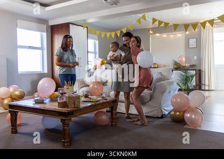 Famiglia diversificata che abbraccia il bambino in piedi accanto a un tavolino da caffè pieno di regali e palloncini nel soggiorno Foto Stock