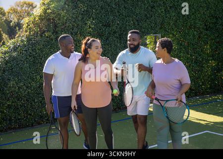 Amici diversi che camminano sul campo da tennis e chiacchierano insieme tenendo racchette e palla da tennis Foto Stock