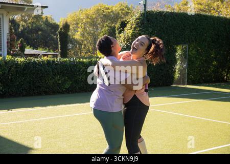 Diverse amiche in abbigliamento sportivo che abbracciano il campo da tennis sul retro con racchette alla luce del sole Foto Stock