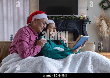 Coppie diverse che si accoccolano sotto una lussuosa coperta sul divano nei cappelli di babbo natale leggendo un libro accanto al caminetto Foto Stock