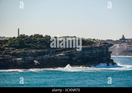 Vista sopraelevata delle scogliere costiere con onde che si infrangono contro di loro e una città lontana sullo sfondo, che mostra la bellezza naturale e l'architetto del mare Foto Stock
