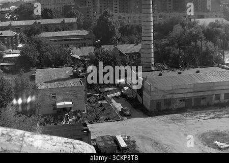Una zona industriale e di servizi in via Iskry (ora via Shelkovichna) a Slovyansk, RSS Ucraina, durante gli anni '1980 Il paesaggio urbano presenta un alto camino in mattoni proveniente da una caldaia locale, circondato da edifici di botteghe, sullo sfondo dei quartieri residenziali della città. Questo scatto riflette il carattere utilitaristico di una città industriale sovietica e simboleggia la vita tranquilla e lavorativa nel Donbas, dove le infrastrutture funzionanti erano la base stessa della stabilità e della routine quotidiana per la comunità Foto Stock