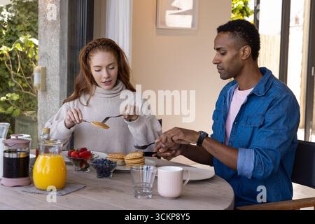 Coppie diverse che mangiano pancake e sorseggiano caffè sul tavolo nell'area da pranzo di casa accanto al giardino Foto Stock