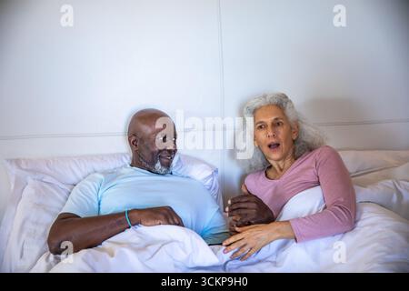Diverse coppie anziane che si tengono per mano e reagiscono con sorpresa nel letto sotto piumone e cuscini bianchi Foto Stock
