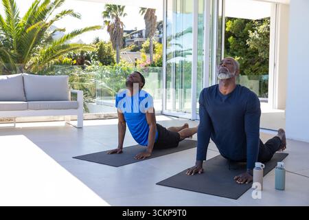 Padre e figlio afroamericani che praticano yoga sulla terrazza soleggiata con stuoie per yoga e bottiglie d'acqua Foto Stock