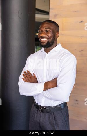 Sorridente uomo afroamericano appoggiato su una colonna scura da un muro di legno, indossando la cintura nella lobby Foto Stock