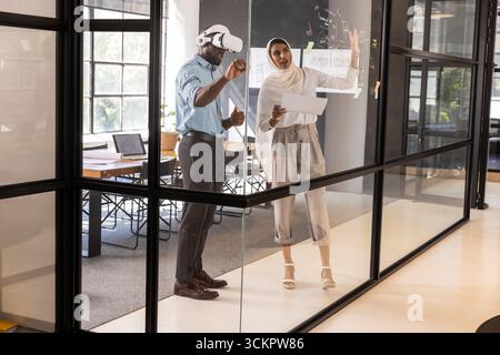 Colleghi diversi in piedi vicino a un pannello di vetro in ufficio, utilizzando attrezzi vr in possesso di piani stampati Foto Stock