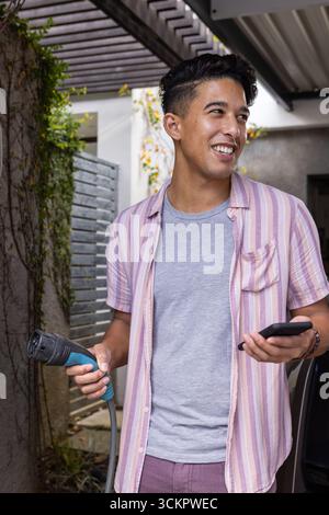 Uomo asiatico che carica un veicolo elettrico in un autoportante tenendo lo smartphone accanto a un muro coperto di vite Foto Stock