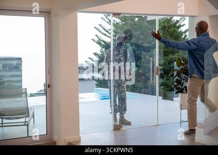 Padre afroamericano che saluta il figlio adulto in uniforme mimetica alla porta sul patio vicino alla piscina Foto Stock