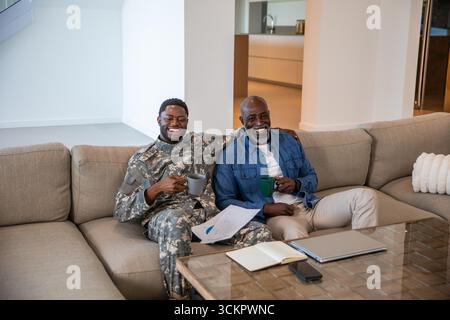 Padre e figlio afroamericani anziani seduti sul divano a casa con tazze da caffè che recensiscono i documenti Foto Stock