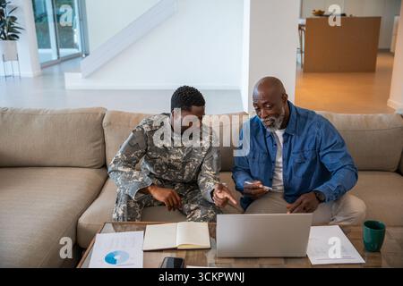 Padre afroamericano senior e figlio soldato che esamina documenti, grafici a casa su un laptop Foto Stock