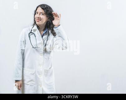 Dottoressa latina che ascolta pettegolezzi, guarda la macchina fotografica. Medico con la mano sull'orecchio ascoltando voci, isolato Foto Stock