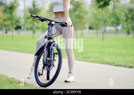 Le gambe femminili sottili indossate da atletica si stagliano accanto alla mountain bike su un sentiero liscio in un parco. Alberi verdi e erba sono visibili sullo sfondo Foto Stock