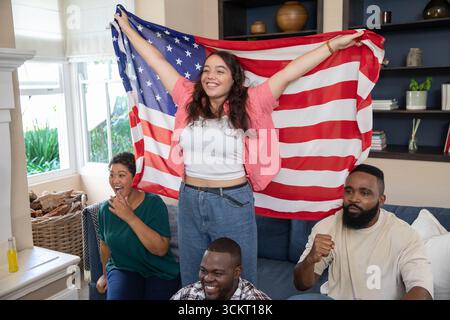 Amici diversi che tifanno il tifo e tengono la bandiera americana sul divano nel soggiorno vicino alla libreria Foto Stock