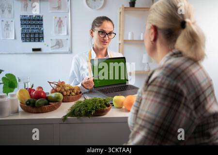 Medico nutrizionista e paziente in sovrappeso che discute qualcosa con schermo verde sul laptop in una clinica alimentare. copia spazio Foto Stock