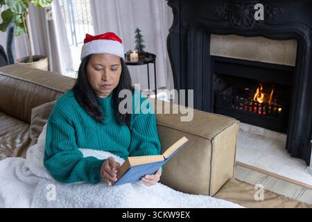 Donna seduta a casa sul divano che legge un libro con copertina rigida sotto una lussuosa coperta con cappello di babbo natale Foto Stock