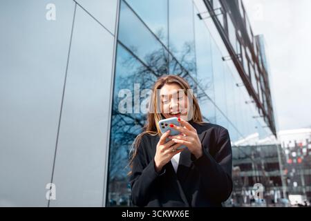 La giovane donna sorride mentre usa il suo smartphone all'aperto in un ambiente urbano con un'architettura moderna che riflette la luce del sole e l'energia vibrante Foto Stock
