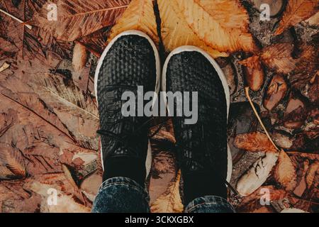 Passeggiata autunnale attraverso le foglie cadute. I piedi di una persona in sneakers nere sono visti camminare su un letto di foglie autunnali cadute, creando un ambiente accogliente e stagionale Foto Stock